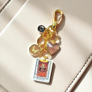 May include: Gold-toned keychain with multiple charms. Charms include a butterfly, a heart, and a tarot card that reads "JUSTICE". The keychain is attached to a gold clasp.