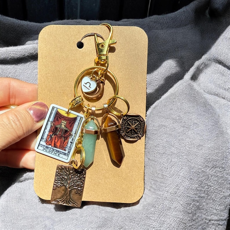 May include: A gold keychain with a variety of charms, including a tarot card, a compass, a crystal, and a tree of life. The tarot card is the Justice card, and the compass is a traditional compass with a star in the center. The crystal is a green crystal, and the tree of life is a small, bronze-colored charm.