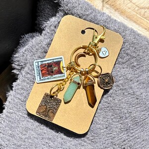 May include: Gold keychain with a variety of charms, including a tarot card, a tree, a compass, and two crystals. The tarot card is the Justice card, and the crystals are green and brown.