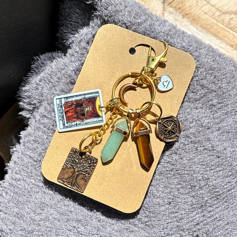 May include: Gold keychain with a variety of charms, including a tarot card, a tree, a compass, and two crystals. The tarot card is the Justice card, and the crystals are green and brown.