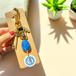 May include: Gold-toned keychain with a key, feather, bell, and blue stone pendant. A white and blue circular charm with a geometric design hangs below the stone. The keychain is attached to a brown card.