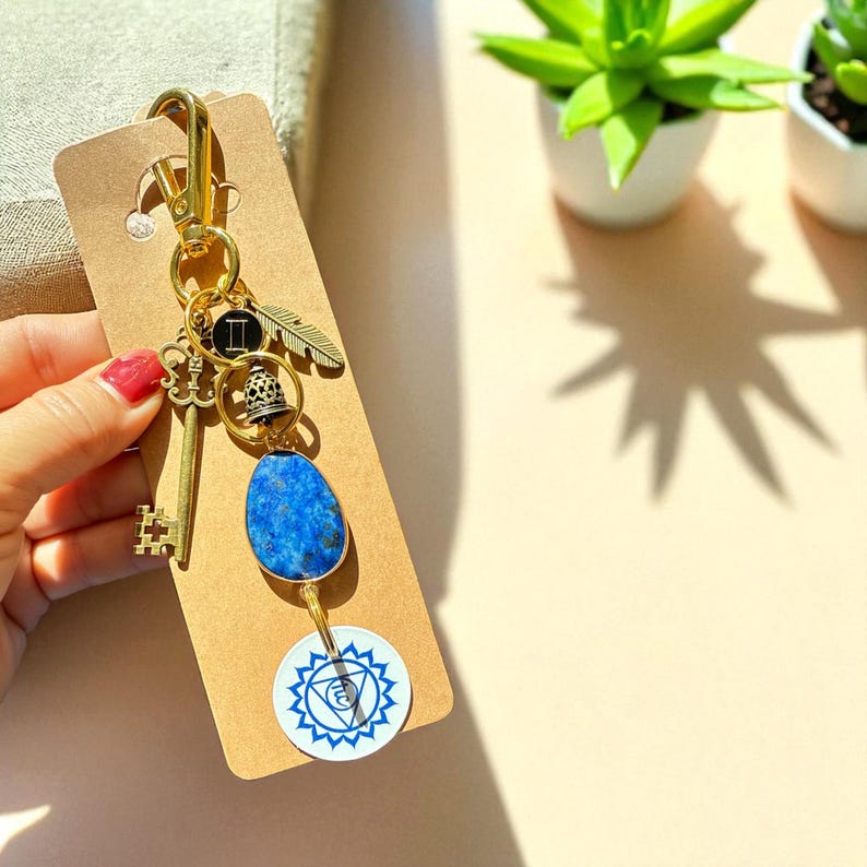 May include: Gold-toned keychain with a key, feather, bell, and blue stone pendant. A white and blue circular charm with a geometric design hangs below the stone. The keychain is attached to a brown card.