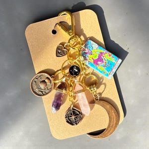 May include: A gold-colored keychain with various charms, including a coin, crystals, a crescent moon, and a tarot card that reads "THE DEVIL". The keychain is attached to a brown card.