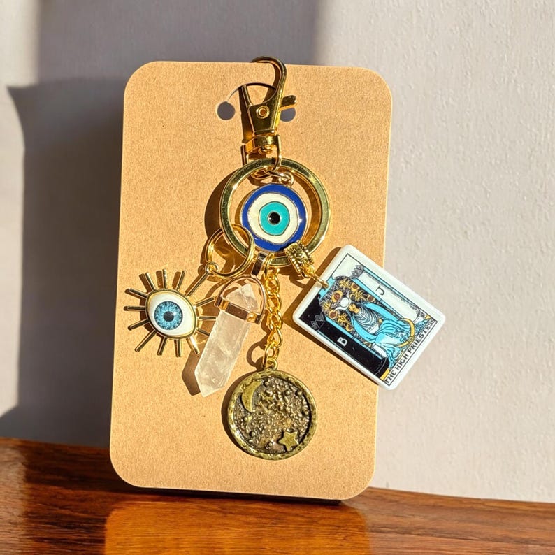 Peut inclure: Porte-cl&eacute;s en or avec un charme &oelig;il bleu mal&eacute;fique, un charme en cristal transparent, un charme lune et &eacute;toile en or et un charme de carte de tarot qui dit "La Grande Pr&ecirc;tresse".