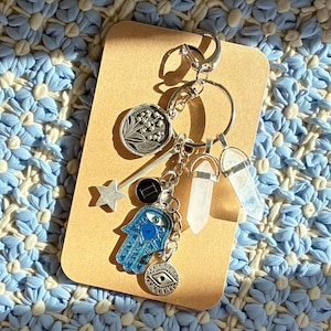 May include: A silver keychain with various charms, including a star, a Hamsa hand, and two crystal pendants. The Hamsa hand is blue with an eye design. The keychain is on a brown card, set against a blue and white floral background.