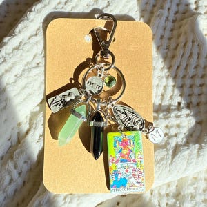 Puede incluir: Un llavero plateado con múltiples dijes, incluyendo un cristal verde, un cristal negro y una pequeña carta de tarot que dice "THE CHARIOT". Otros dijes incluyen un helecho, un diseño de fases lunares y la letra "M".