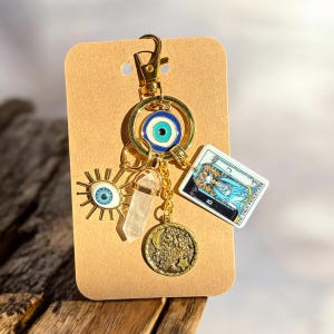 Peut inclure: Porte-cl&eacute;s en or avec un charme &oelig;il bleu mal&eacute;fique, un charme en cristal transparent, un charme lune et &eacute;toile en or et un charme de carte de tarot avec le texte "La Grande Pr&ecirc;tresse".