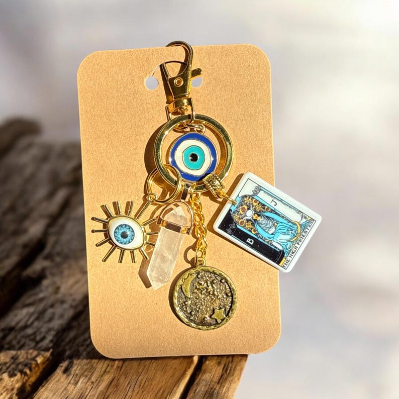 Peut inclure: Porte-cl&eacute;s en or avec un charme &oelig;il bleu mal&eacute;fique, un charme en cristal transparent, un charme lune et &eacute;toile en or et un charme de carte de tarot avec le texte "La Grande Pr&ecirc;tresse".