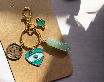 May include: Gold key chain with a green crystal, a heart charm with an evil eye, a gold charm with a crescent moon and sun, and a green planet charm.