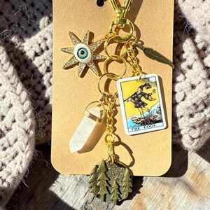 May include: A gold-toned keychain featuring multiple charms attached to a central ring: a star-shaped eye charm with a green iris, a clear crystal point, a rectangular tarot card charm labeled The Fool, a small feather, and a round tree-etched disc.