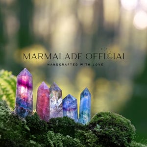 May include: Five colorful crystal points in shades of purple, pink, and blue are displayed on a bed of green moss. The crystals are arranged in a row, with the text "MARMALADE OFFICIAL HANDCRAFTED WITH LOVE" above them.