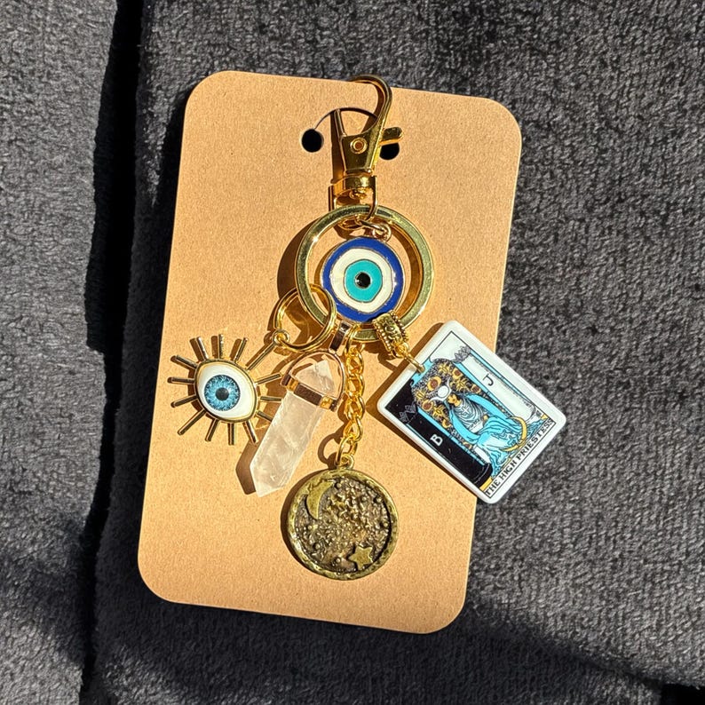 Peut inclure: Porte-cl&eacute;s avec un charme en or, un &oelig;il bleu mal&eacute;fique, un cristal transparent, une carte de tarot avec le texte "La Grande Pr&ecirc;tresse" et un charme de lune en or.