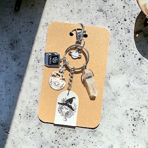 May include: A silver keychain with a variety of charms, including a book with the word "Spells" on it, a bottle with a crescent moon and eye, a crystal, and a witch with a crescent moon on her hat.