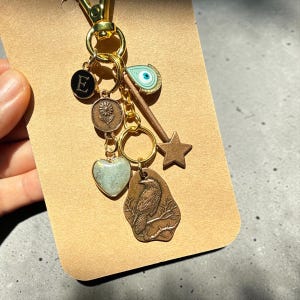 Op de afbeelding: Een goudkleurige metalen sleutelhanger met diverse bedels: een zwarte ronde bedel met de letter E, een bronzen zonnebloem munt, een hartvormige bedel, een lichtblauw boze oog, een bronzen ster en een bronzen bedel van een raaf.