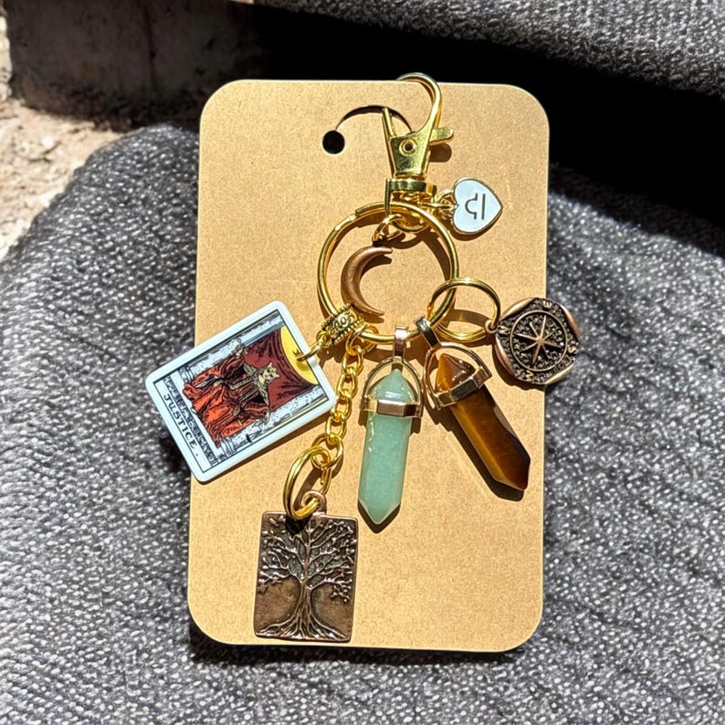 May include: Gold keychain with a variety of charms, including a tarot card, a tree of life, a compass, and crystals. The tarot card is the Justice card.