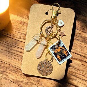 May include: A gold-colored keychain with multiple charms. The charms include two crystal pendants, a tarot card, a tree of life pendant, a star, and a heart. The tarot card reads "THE DEVIL". The keychain is attached to a brown card.