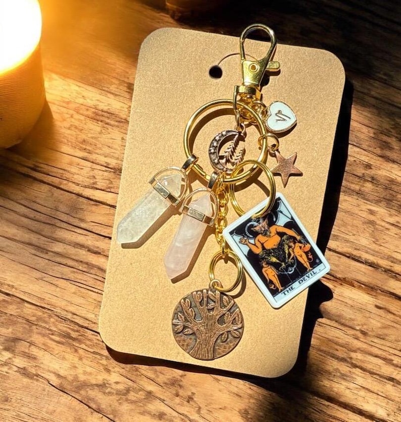 May include: A gold-colored keychain with multiple charms. The charms include two crystal pendants, a tarot card, a tree of life pendant, a star, and a heart. The tarot card reads "THE DEVIL". The keychain is attached to a brown card.