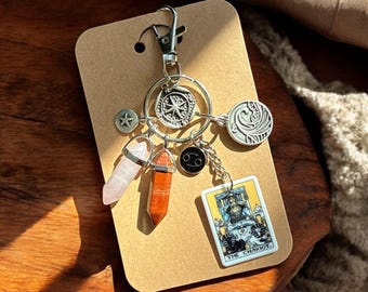 May include: A silver charm keychain with a variety of charms, including a star, a compass, a crystal, a tarot card, and a zodiac symbol. The keychain is on a brown background.