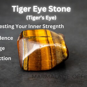 May include: A tiger's eye stone, a brown and gold gemstone with a striated pattern, is shown on a dark gray surface. The text "Tiger Eye Stone (Tiger's Eye)" is above the stone. The text "Manifesting Your Inner Strength" is below the stone. The text "Confidence, Courage, Protection" is below the text "Manifesting Your Inner Strength". The text "MARMALADE OFFICIAL" is below the stone.