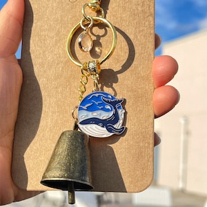 May include: Gold keychain with a blue whale charm, a small bell, and a clear crystal. The whale charm is on a white background with blue waves and flying birds.