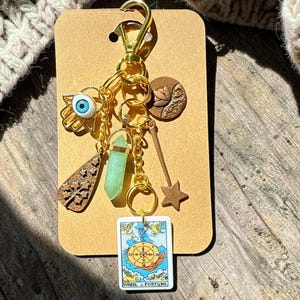 May include: A gold-toned keychain featuring a hamsa hand charm with a blue eye, a light green crystal point, a brown textured tree charm, a brown moon charm, a brown star wand, and a rectangular Wheel of Fortune tarot card charm.