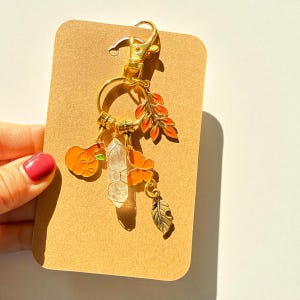 Puede incluir: Un llavero dorado con colgantes con temática otoñal. Los colgantes incluyen una calabaza naranja, un cristal transparente, hojas naranjas, una flor y una pluma. El llavero está sujeto a una tarjeta marrón.