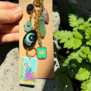 May include: A gold-toned keychain featuring multiple charms including a rectangular Strength tarot card, a green crystal point, a black moon disc, a turquoise hamsa hand, a small Leo zodiac symbol, and a square charm with a central eye design.