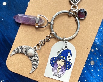 Può includere: Un portachiavi color argento con una punta di cristallo viola, un ciondolo a mezzaluna con fasi lunari e un ciondolo rettangolare bianco con il disegno di una persona che indossa un cappello da strega blu con stelle e pipistrelli, e una perlina.