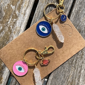 Op de afbeelding: Twee gouden sleutelhangers met blauwe en roze boze oogcharms, heldere kwartskristallen en gouden accenten. De ene sleutelhanger heeft een halve maan charme en de andere heeft een rode planeet charme.