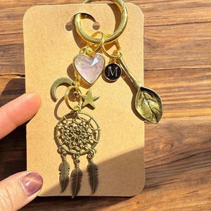 Puede incluir: Llavero dorado con una variedad de dijes, incluyendo un corazón rosa, una luna creciente, una estrella, un atrapasueños y una hoja. El llavero también tiene un pequeño dije con la letra "M" en él.