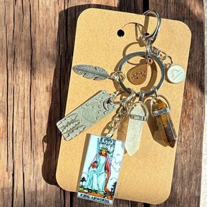May include: A silver keychain with multiple charms, including a feather, a mountain scene, a tarot card with the text "King of Swords", a clear crystal, and a brown crystal. The keychain is attached to a brown card.