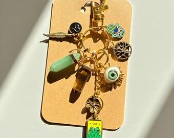 May include: A gold-colored keychain with various charms, including a green crystal, a tiger's eye crystal, a Hamsa hand, an eye, a feather, a tree of life, and a tarot card. The charms are attached to a gold clasp and a key ring.