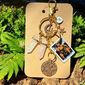 May include: A gold-colored keychain with various charms, including two crystal pendants, a tarot card, a star, a heart, a crescent moon, and a tree of life pendant. The tarot card depicts a devil figure. The keychain is attached to a brown cardboard backing.