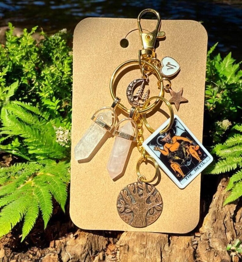 May include: A gold-colored keychain with various charms, including two crystal pendants, a tarot card, a star, a heart, a crescent moon, and a tree of life pendant. The tarot card depicts a devil figure. The keychain is attached to a brown cardboard backing.