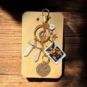 May include: A gold-colored keychain with various charms, including two crystal pendants, a tarot card, a star, a heart, and a tree of life pendant. The tarot card depicts a devil figure.