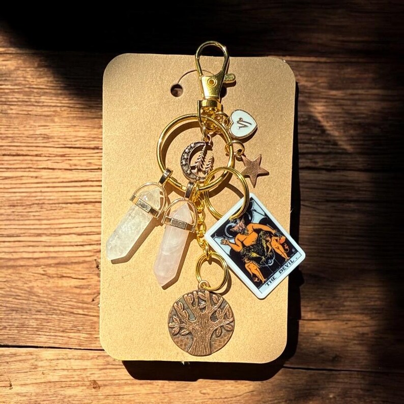 May include: A gold-colored keychain with various charms, including two crystal pendants, a tarot card, a star, a heart, and a tree of life pendant. The tarot card depicts a devil figure.