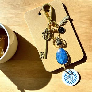 May include: A gold-colored keychain with various charms, including a key, feather, bell, and a blue stone pendant. A white circular charm with a blue design is also attached. The keychain is displayed on a brown card.
