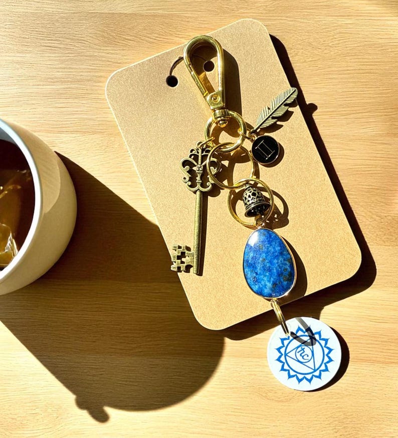 May include: A gold-colored keychain with various charms, including a key, feather, bell, and a blue stone pendant. A white circular charm with a blue design is also attached. The keychain is displayed on a brown card.