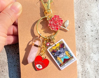 May include: Gold keychain with a pink crystal, a red heart with an evil eye, a gold charm with an evil eye, and a tarot card charm that says "The Lovers".