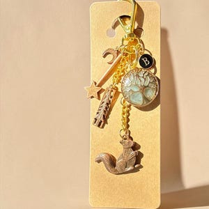 Puede incluir: Un llavero dorado con múltiples colgantes, incluyendo una ardilla, un árbol de la vida, una estrella y una luna creciente. El llavero está sujeto a una etiqueta de cartón marrón.