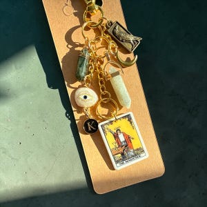 May include: A gold-toned keychain with multiple charms, including crystals, an eye pendant, a letter "K", a bird, a crescent moon, and a tarot card. The tarot card features an illustration of a magician. The keychain is attached to a brown tag.