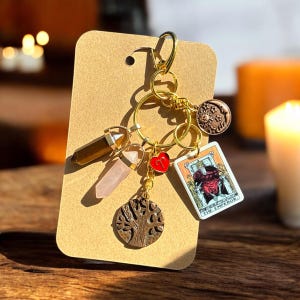 May include: A gold-colored keychain with multiple charms, including a brown crystal, a pink crystal, a red heart, a tree of life pendant, a sun-shaped charm, and a tarot card that reads "The Emperor". The keychain is attached to a brown card.