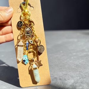 May include: A gold-colored keychain with multiple charms, including crystals, a blue hand with an eye, and a coin. The keychain is attached to a brown card. The charms are various shapes and sizes, with gold and blue accents.