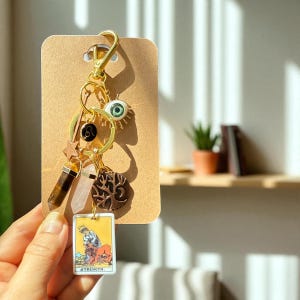 Op de afbeelding: Een goudkleurige sleutelhanger met diverse bedels, waaronder een groen oog, een ster, een boom en een tarotkaart met de tekst "Strength". De sleutelhanger heeft ook twee kristallen en een dierenriemteken. De sleutelhanger is bevestigd aan een bruine kaart.