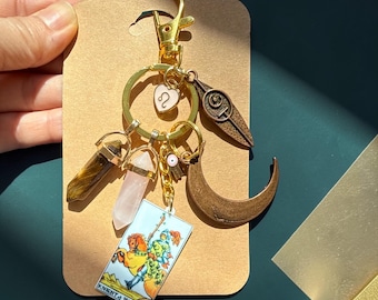 May include: A gold-colored keychain with various charms, including a tiger's eye crystal, a rose quartz crystal, a tarot card, a crescent moon, and a heart-shaped charm. The tarot card reads "Knight of Wands."
