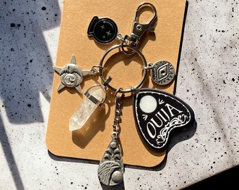 May include: A silver charm keychain with a variety of charms, including a crystal, a moon, a star, an eye, a cauldron, and a black and white Ouija board charm.