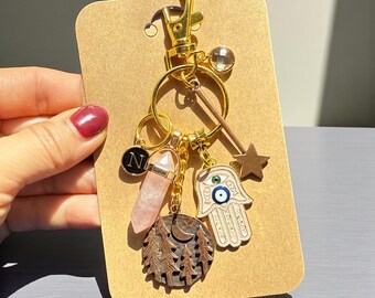 May include: A gold-colored keychain with multiple charms. Charms include a pink crystal, a Hamsa hand with an evil eye, a star, a forest scene, and a letter "N" pendant. The keychain is attached to a gold clasp.