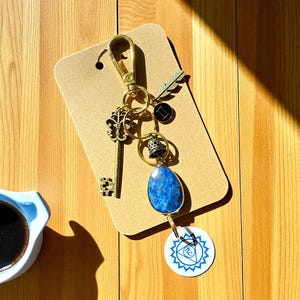 May include: A gold-colored keychain with various charms, including a key, a feather, and a blue stone pendant. The keychain is attached to a brown tag and rests on a wooden surface. A cup of coffee is in the lower left corner.