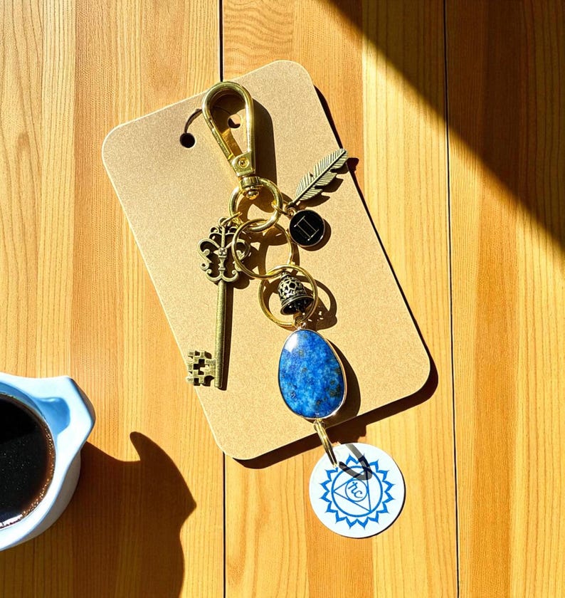 May include: A gold-colored keychain with various charms, including a key, a feather, and a blue stone pendant. The keychain is attached to a brown tag and rests on a wooden surface. A cup of coffee is in the lower left corner.
