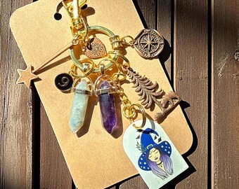 Può includere: Un portachiavi dorato con vari ciondoli, tra cui una stella, cristalli, una bussola e un'illustrazione di una strega. I cristalli sono azzurri e viola. L'illustrazione della strega ha un cappello blu e capelli viola.